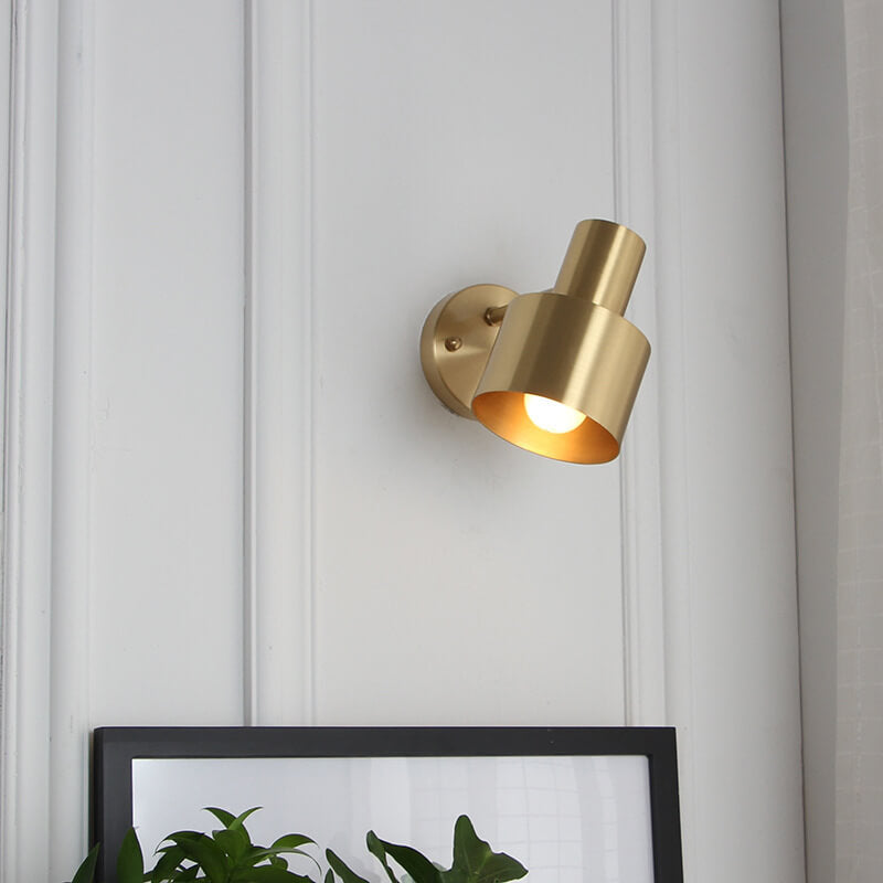 Gold wall-mounted light fixture on a white paneled wall with a plant in the foreground.