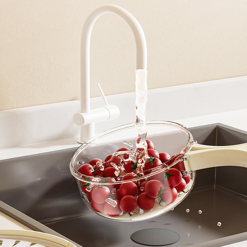 Tomatoes being washed in a glass bowl under running water from a white faucet in a kitchen sink.
