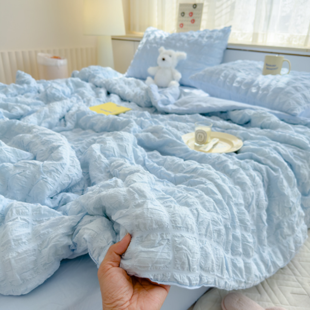 Light blue textured comforter on a bed with a teddy bear and small pillow.