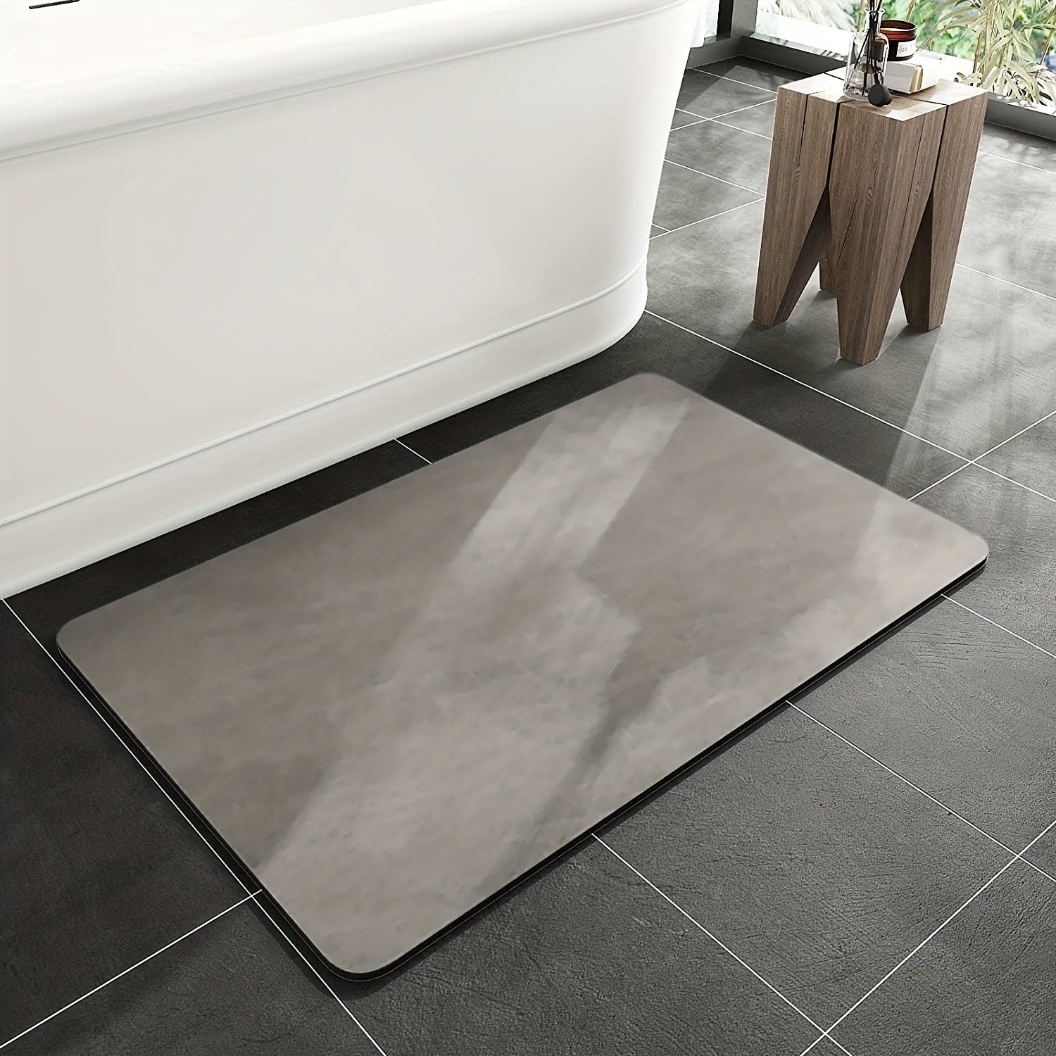 Gray bath mat on a tiled bathroom floor with a bathtub and small table in the background.