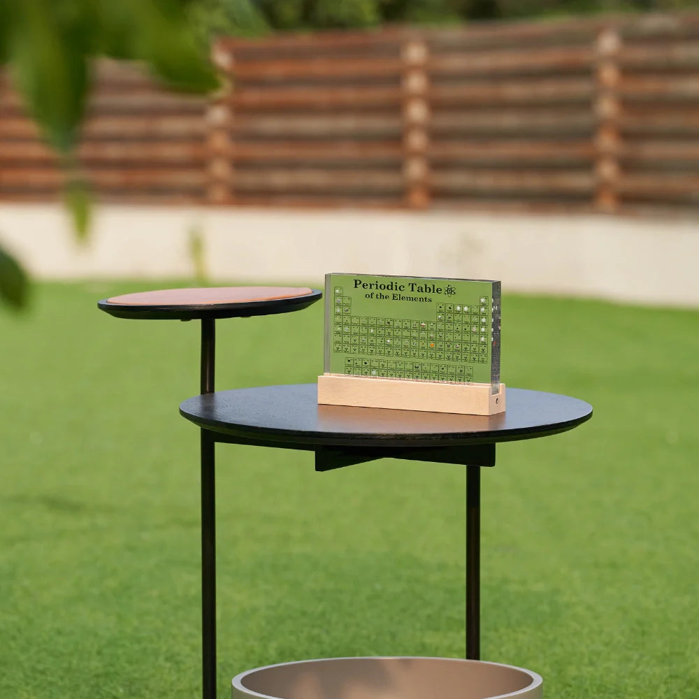 Periodic table on a small round table outdoors with green grass and wooden bench in the background
