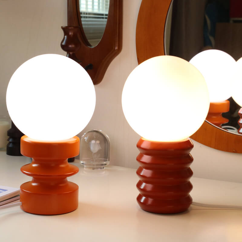 Two spherical lamps with orange bases on a surface, with a mirror and glass object in the background.