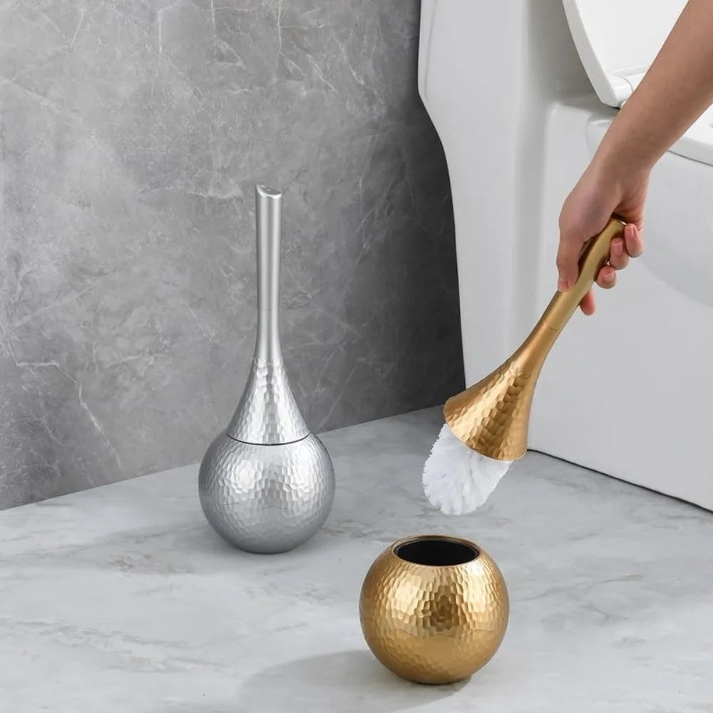 Gold cleaning brush with white bristle head being used on a gold container next to a silver container in a bathroom setting.