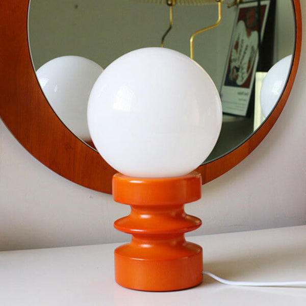 Table lamp with white globe shade and orange base on a surface, with a mirror in the background.