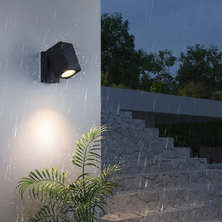 Outdoor wall light fixture on a building exterior during rain with plants and steps in the background.