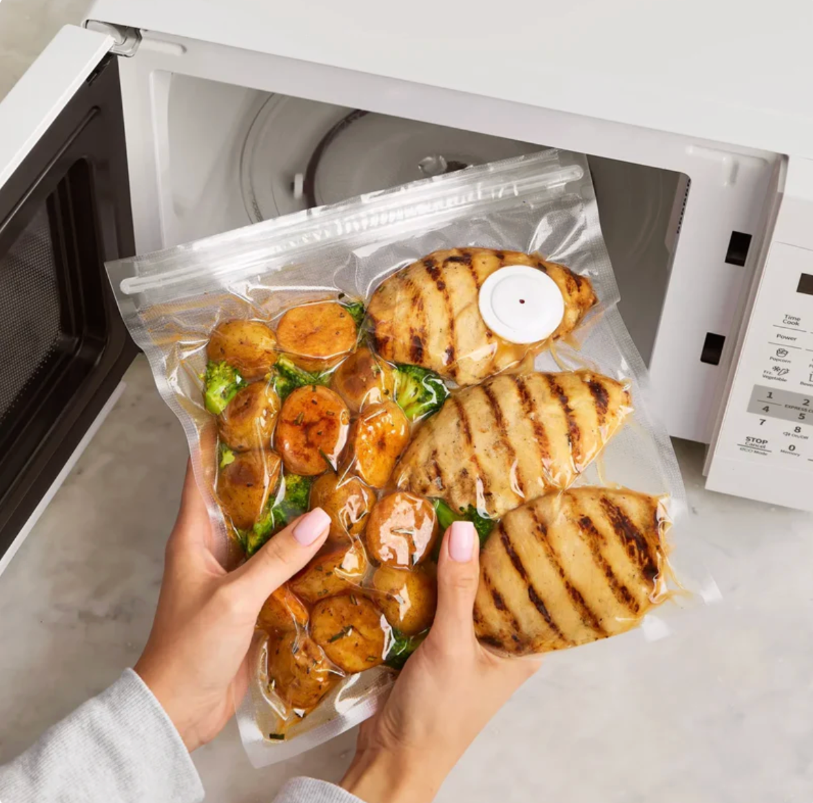 Hand holding a vacuum-sealed package of grilled chicken and vegetables in front of an open microwave.