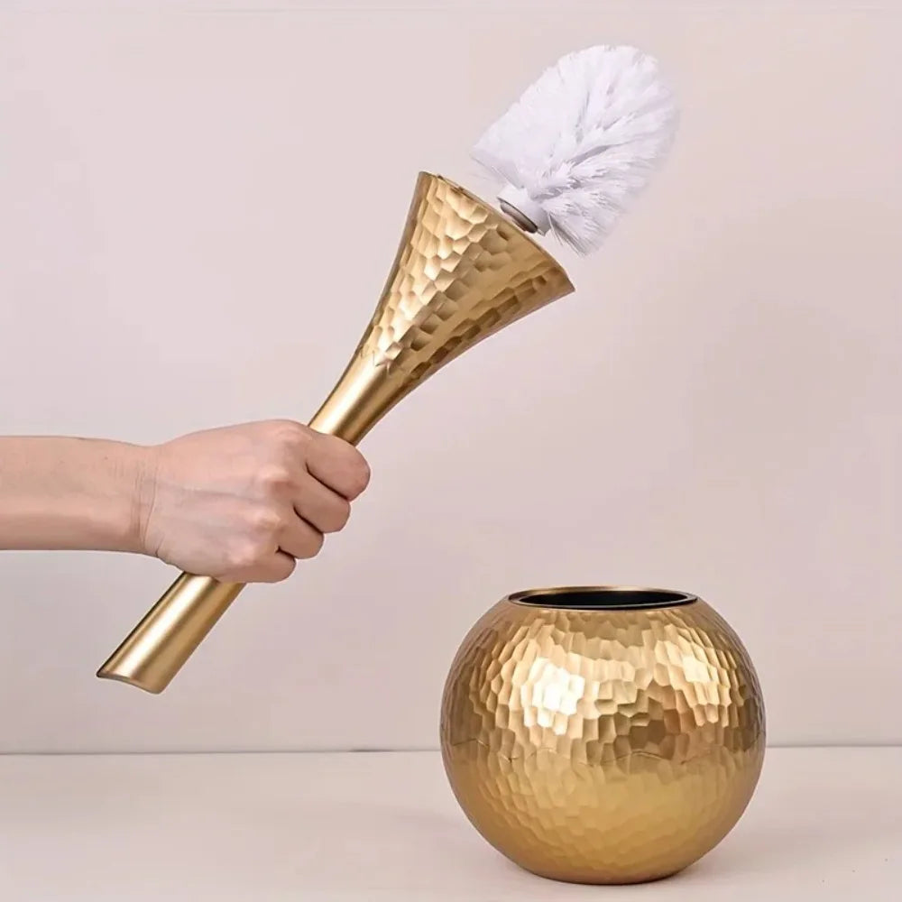 Gold cleaning brush with white bristle head being held above a gold vase on a plain background