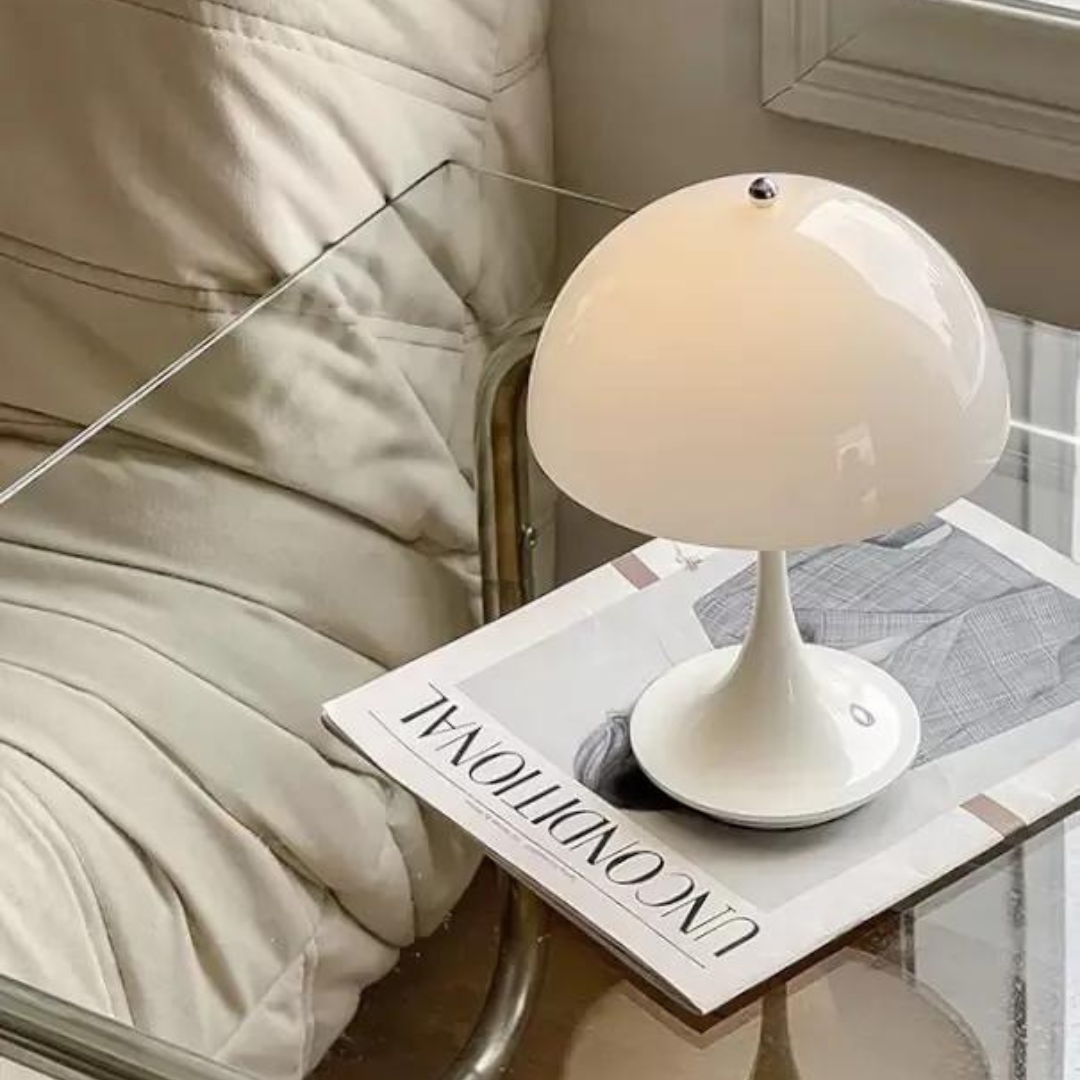 White table lamp on a glass table with a magazine underneath, next to a beige armchair.