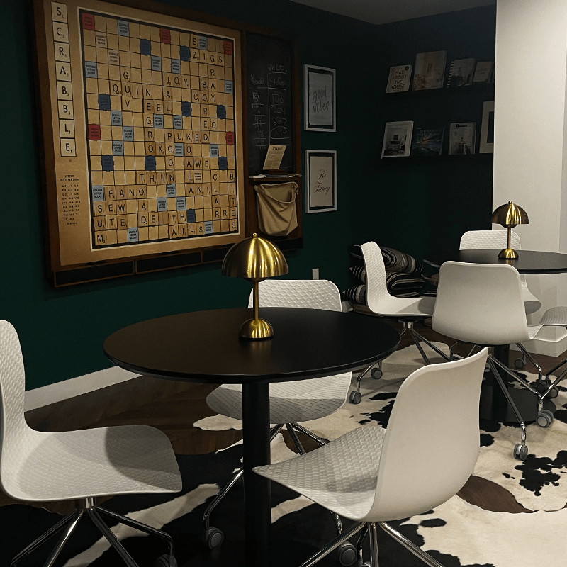 Modern dining room with white chairs, black tables, and a Scrabble board on the wall.