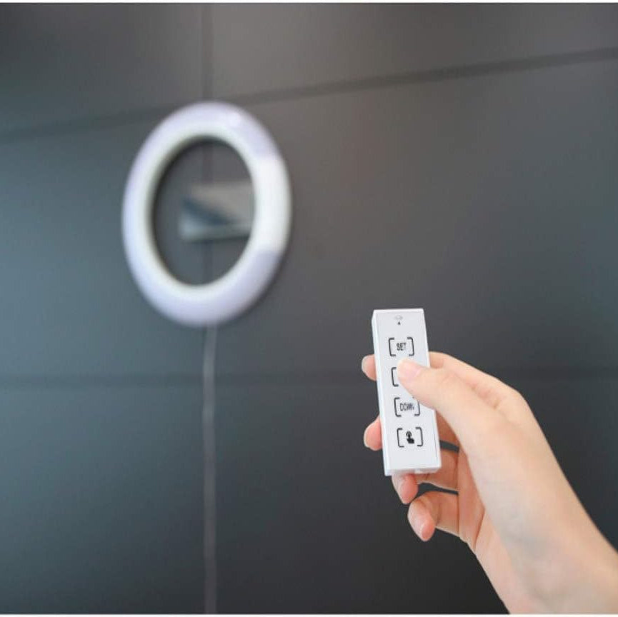 Hand holding a remote control near a circular device on a gray wall.