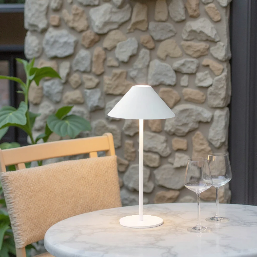 White table lamp on a round marble table with two wine glasses against a stone wall.