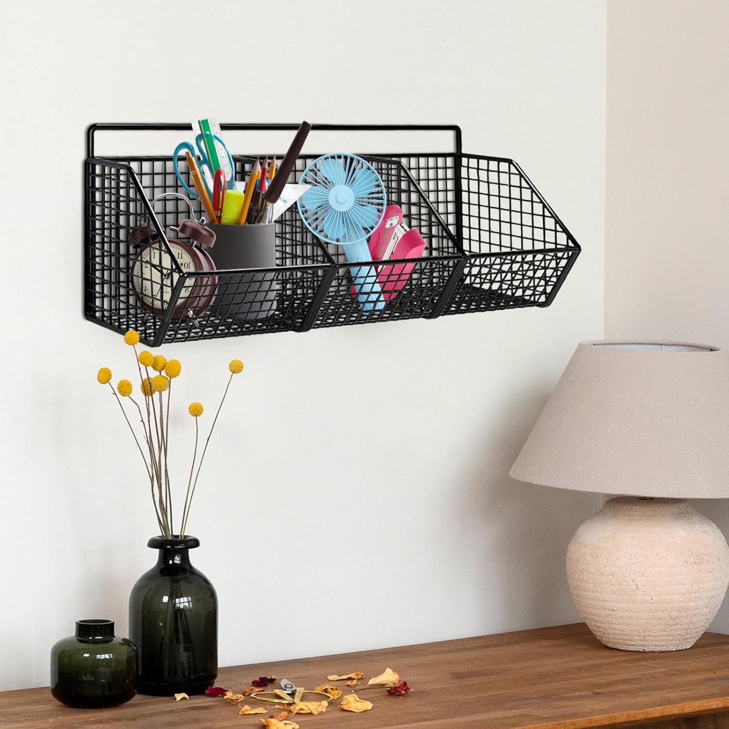 Black metal wall-mounted basket with stationery items on a wooden surface with decor elements.