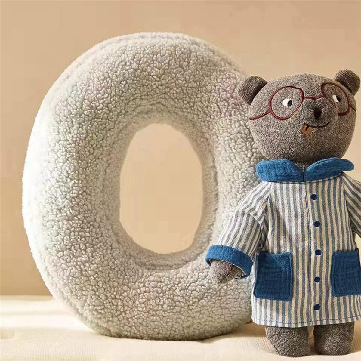 Stuffed bear toy with glasses and a striped coat standing next to a large letter 'O' made of textured material.