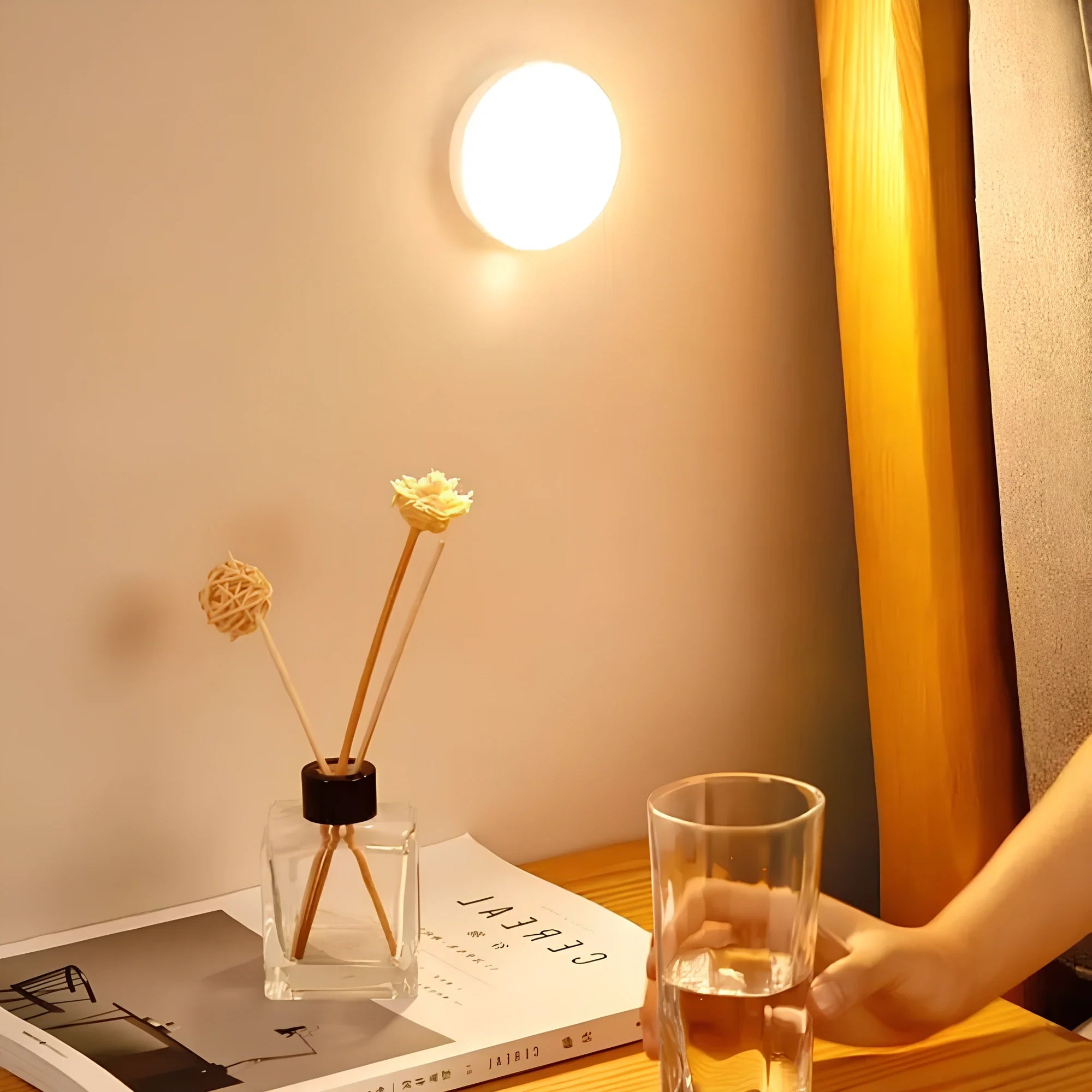 Glass of water on a table next to a diffuser and books, with a light on the wall.