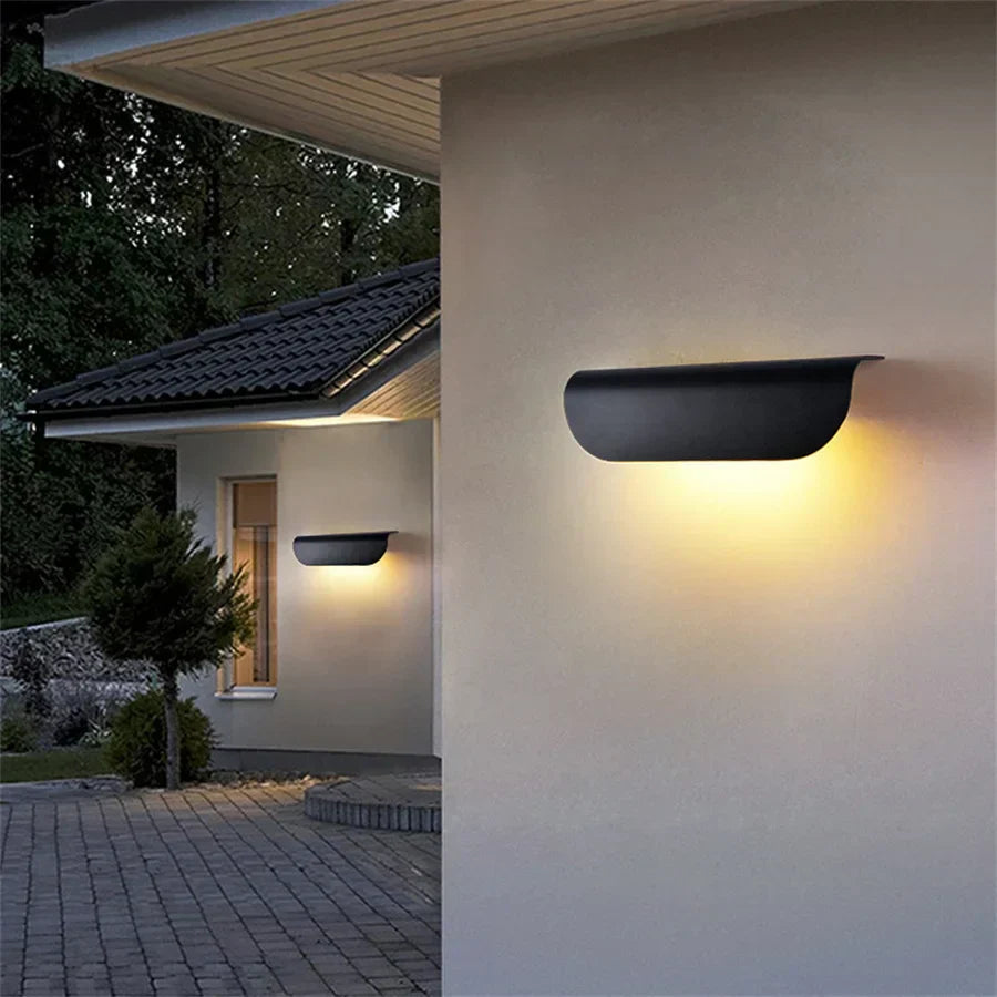Modern outdoor wall lights on a house exterior with a dark roof and greenery.