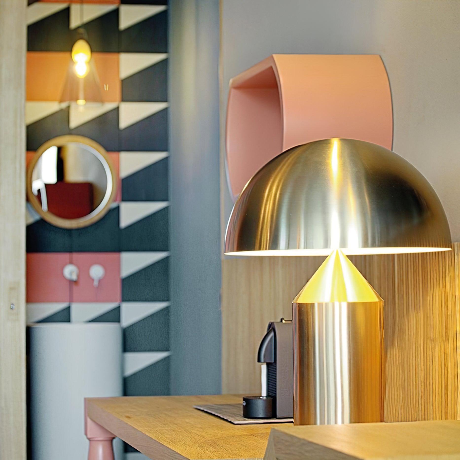 Modern gold table lamp on a wooden desk with a colorful geometric wall design in the background.