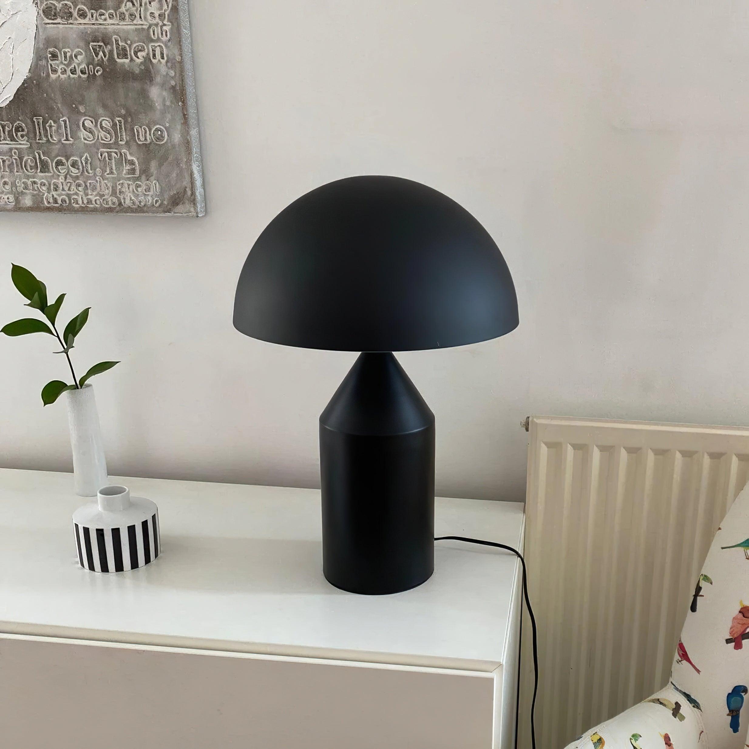 Black table lamp on a white surface with a plant and decorative items in the background.