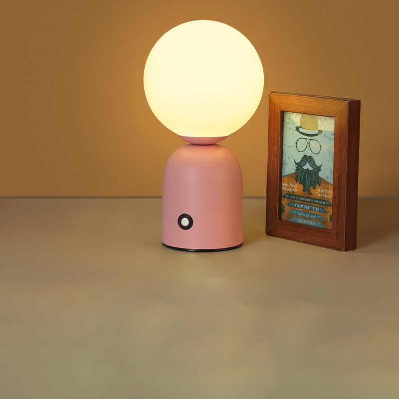 Pink table lamp with a round white bulb next to a wooden-framed picture on a beige surface.