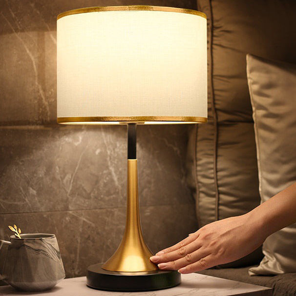Table lamp with gold base and white shade being adjusted by a hand on a marble surface.
