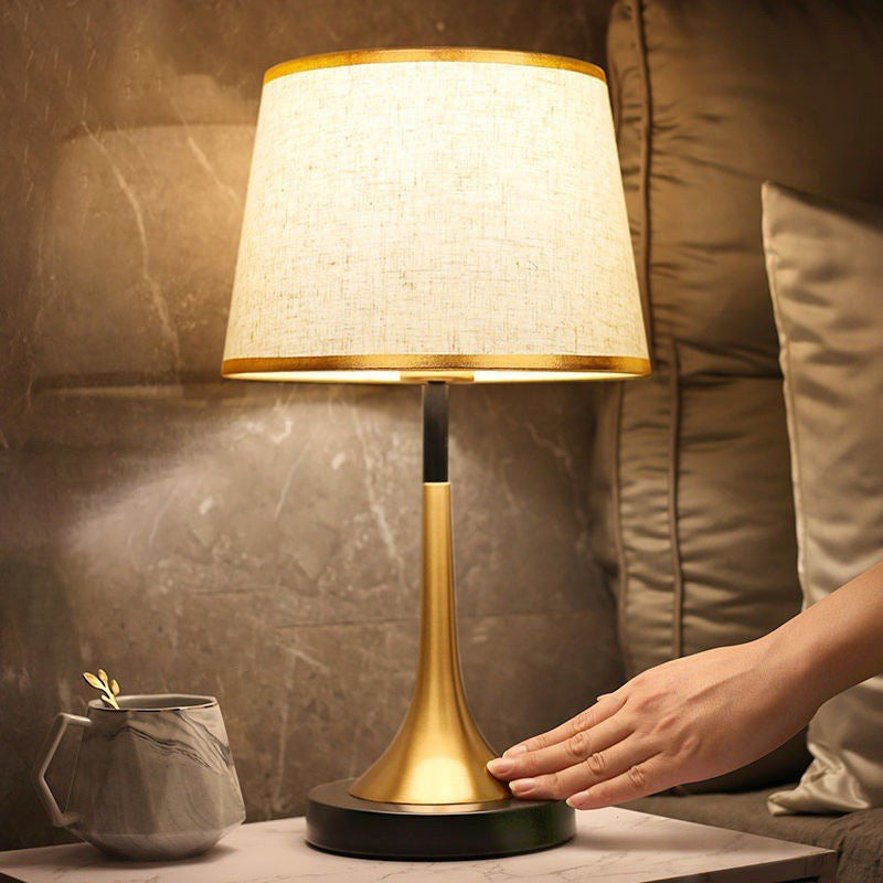 Table lamp with gold base and white shade on a table, hand reaching towards it.
