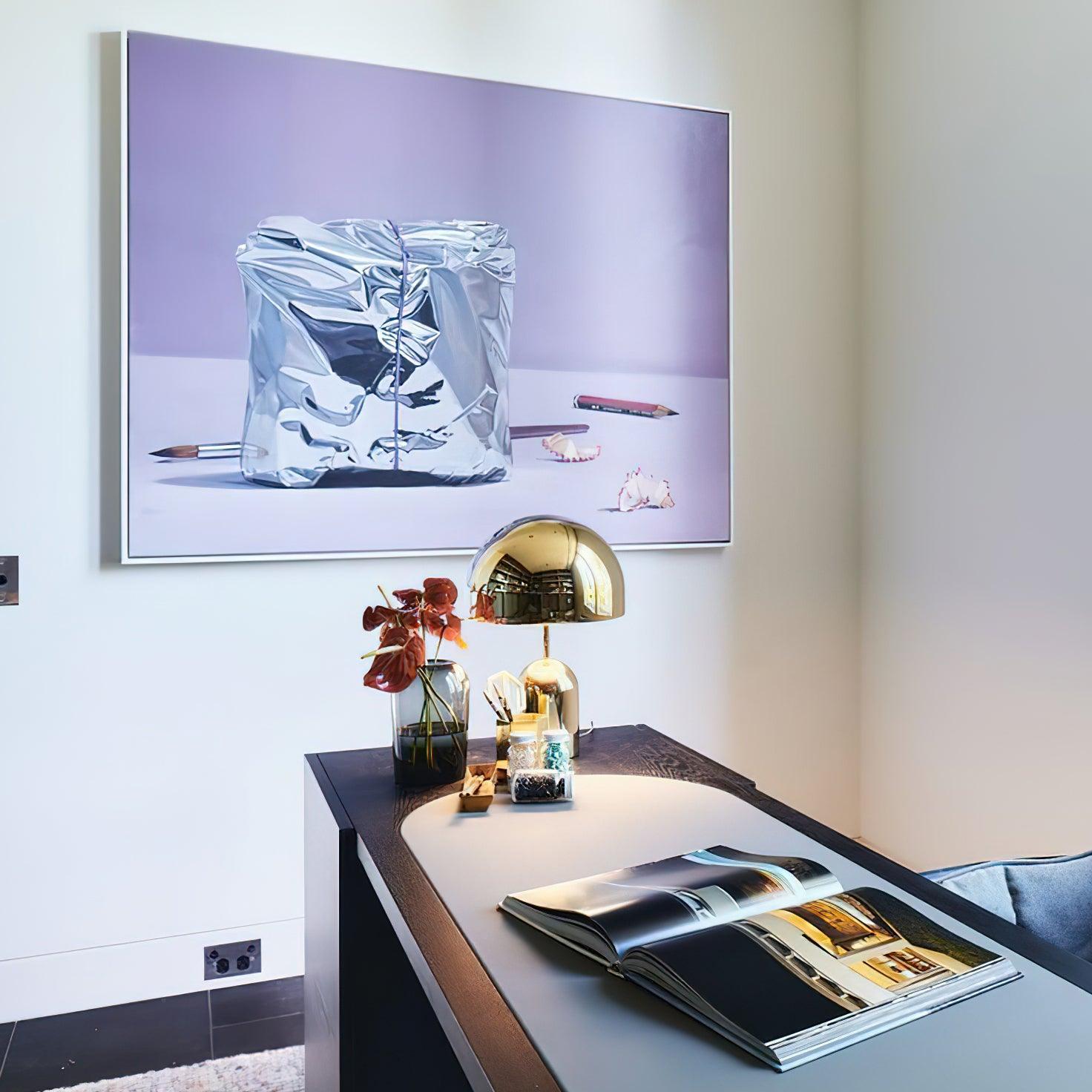 Modern office desk with a lamp, books, and decorative items, featuring a large framed artwork on the wall.