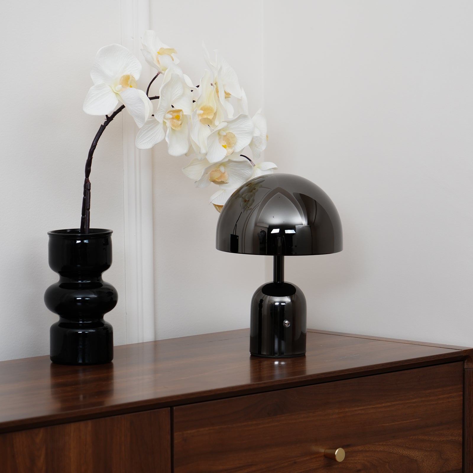 Black table lamp and vase with white flowers on a wooden surface against a light wall.