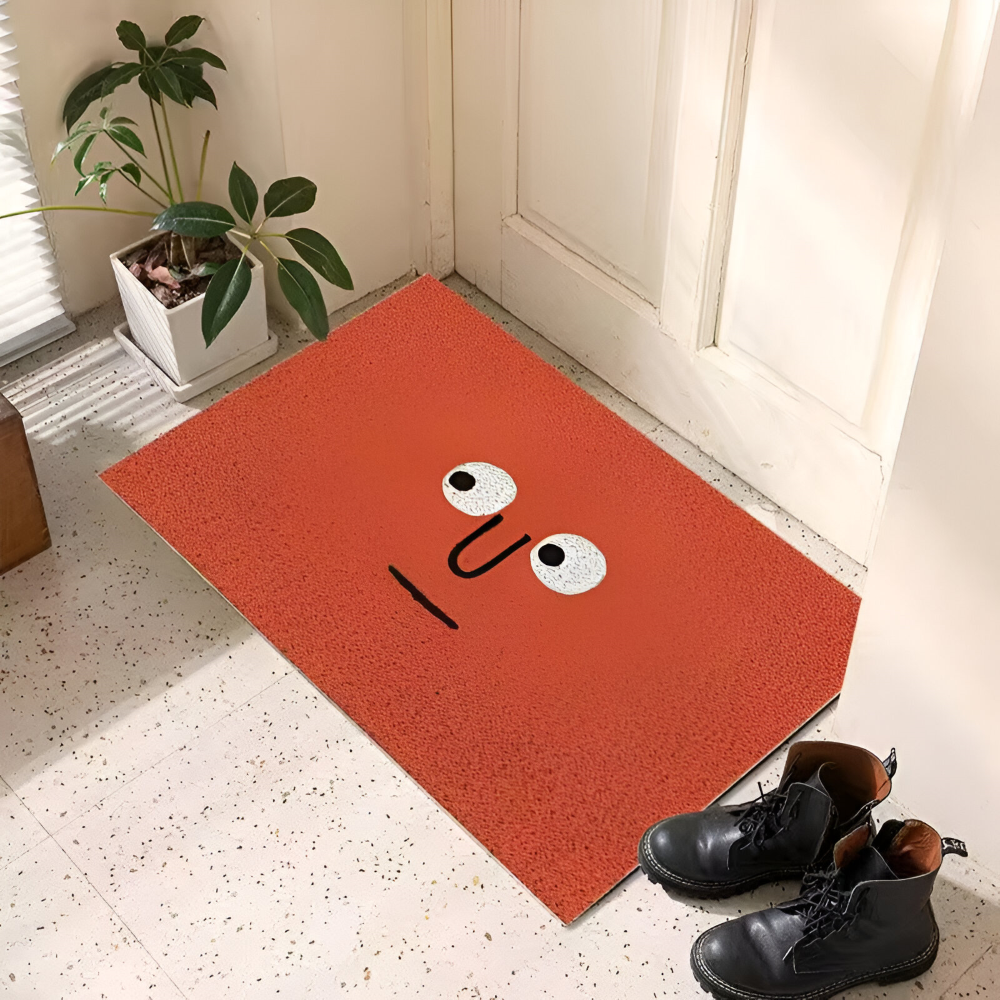 Orange doormat with a face design on a tiled floor next to a plant and shoes.