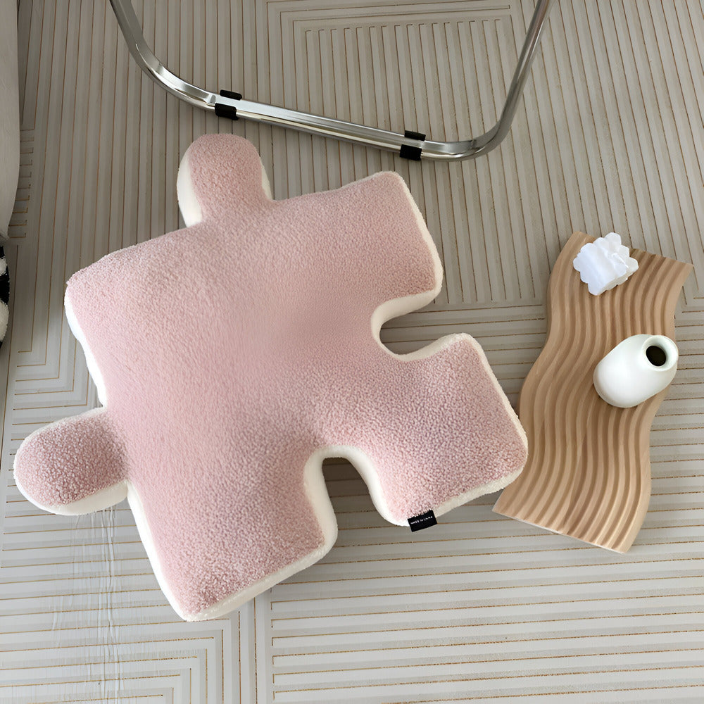 Pink puzzle piece-shaped pillow on a textured surface with a small white vase and decorative item.