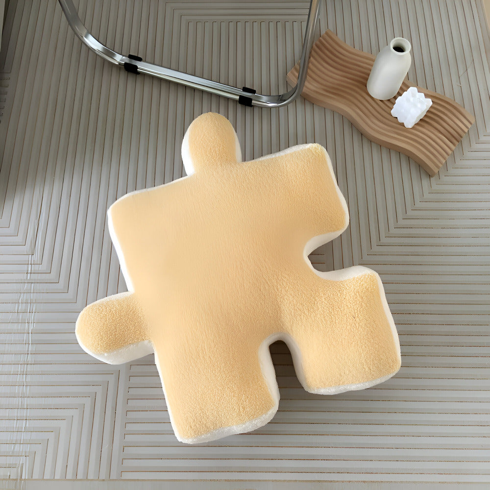 Beige puzzle piece-shaped pillow on a textured surface with a vase and towel in the background.