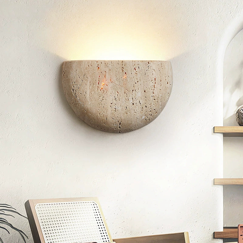 Round stone wall light fixture on a white wall with a chair and shelf in the foreground.