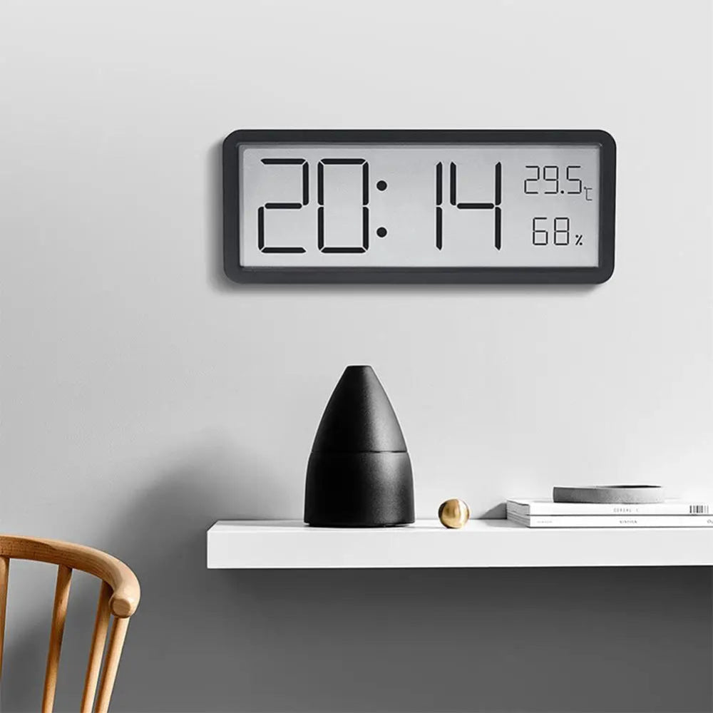 Digital wall clock on a gray wall with a shelf below displaying a black vase and books.