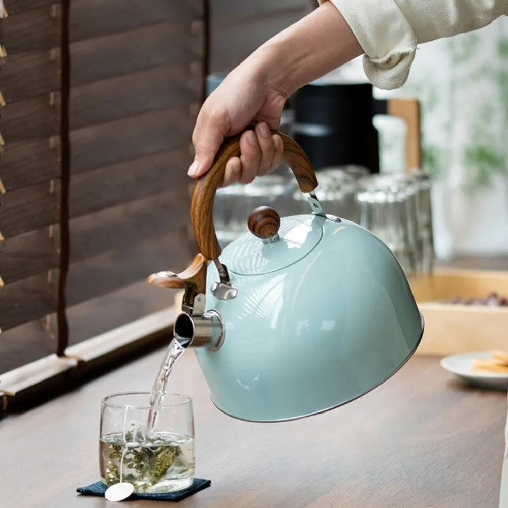 Tea being poured from a light blue kettle with a wooden handle into a glass on a table.