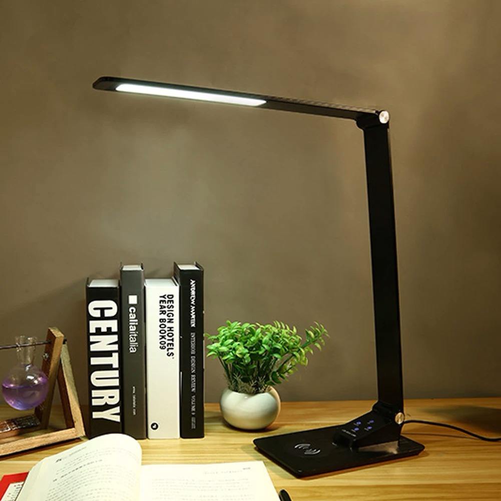 Black LED desk lamp on a wooden surface with books and a plant in the background.