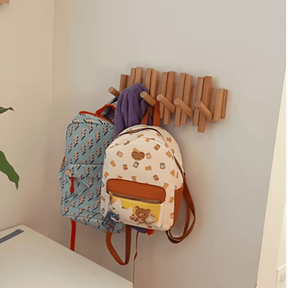 Wooden wall hook with two backpacks hanging on it against a light-colored wall.