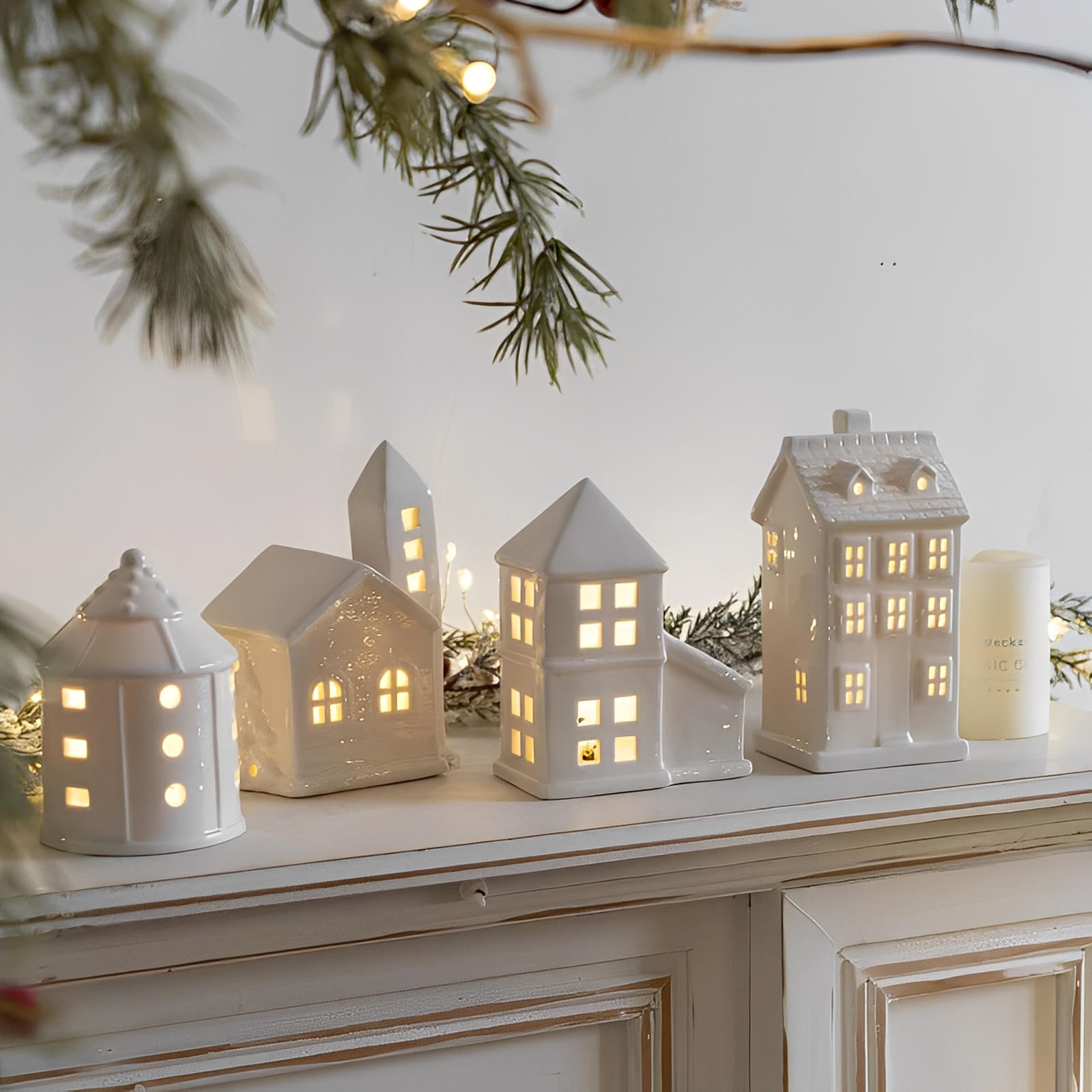 Set of white ceramic house-shaped lanterns on a mantelpiece with Christmas decorations.
