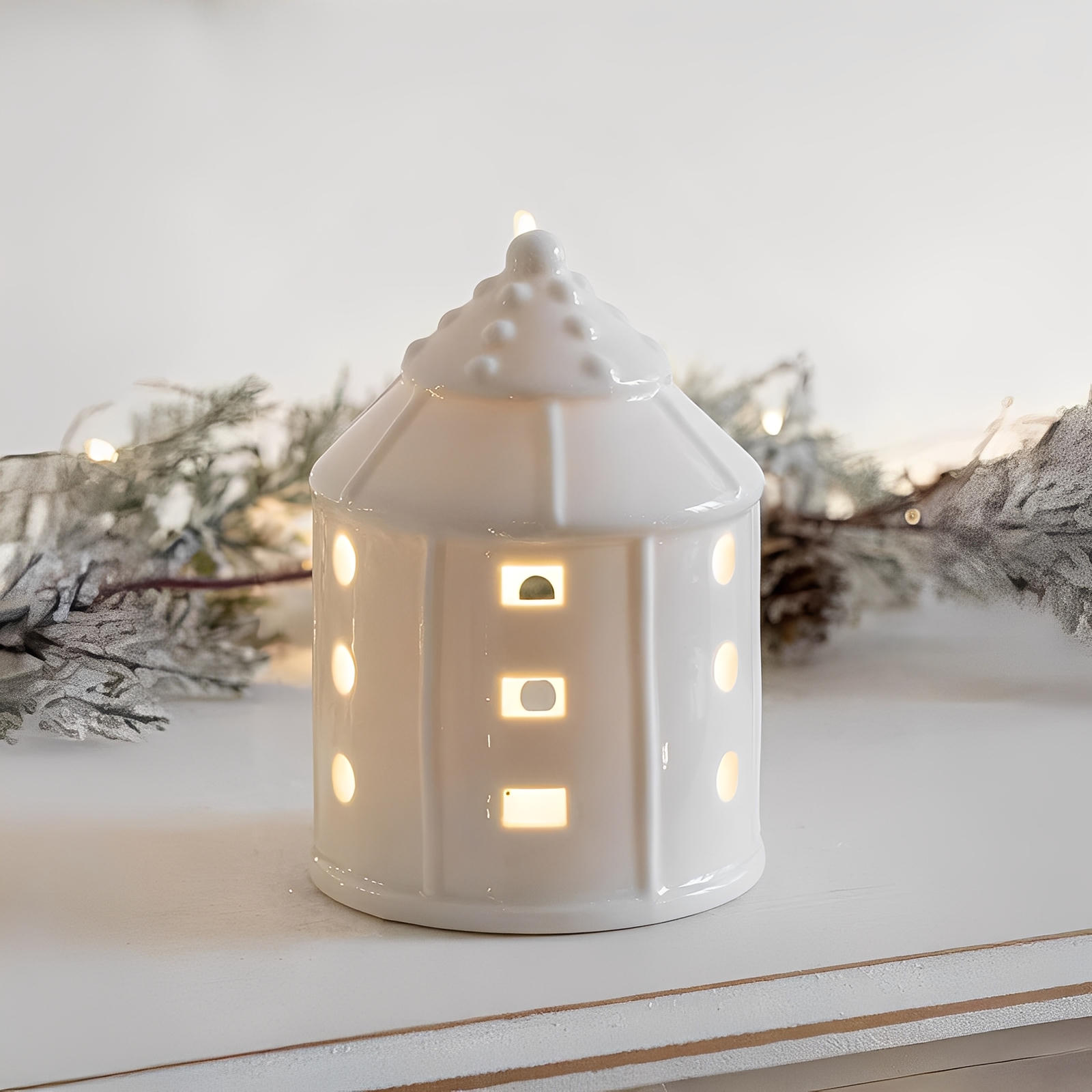 White ceramic house-shaped lantern with lit interior on a snowy surface with evergreen branches.