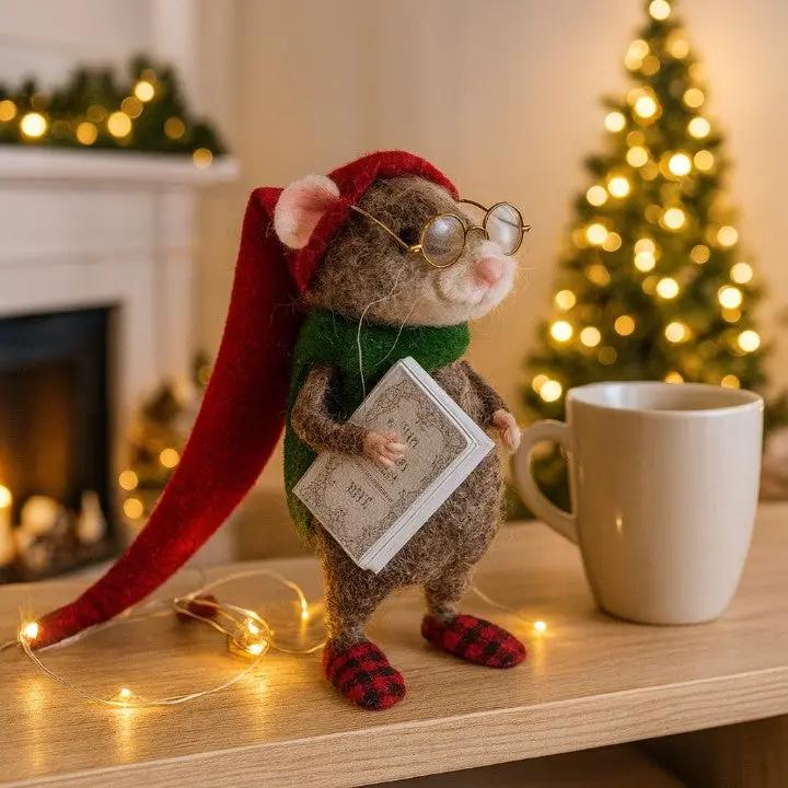 Stuffed mouse toy with a red hat and glasses holding a book, next to a mug on a table with Christmas decorations in the background.