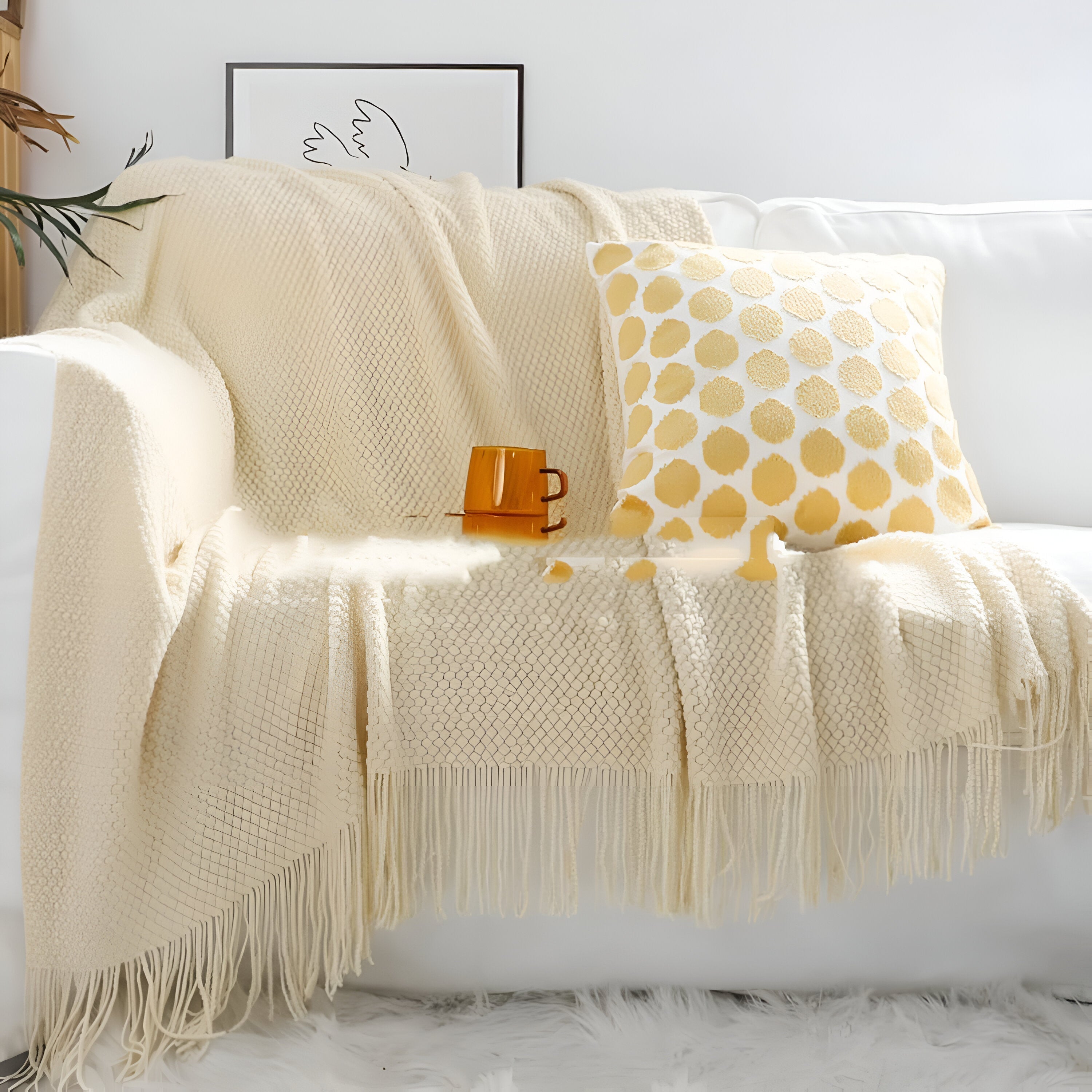 Beige throw blanket draped over a sofa with a yellow polka dot pillow and a cup on a white surface.
