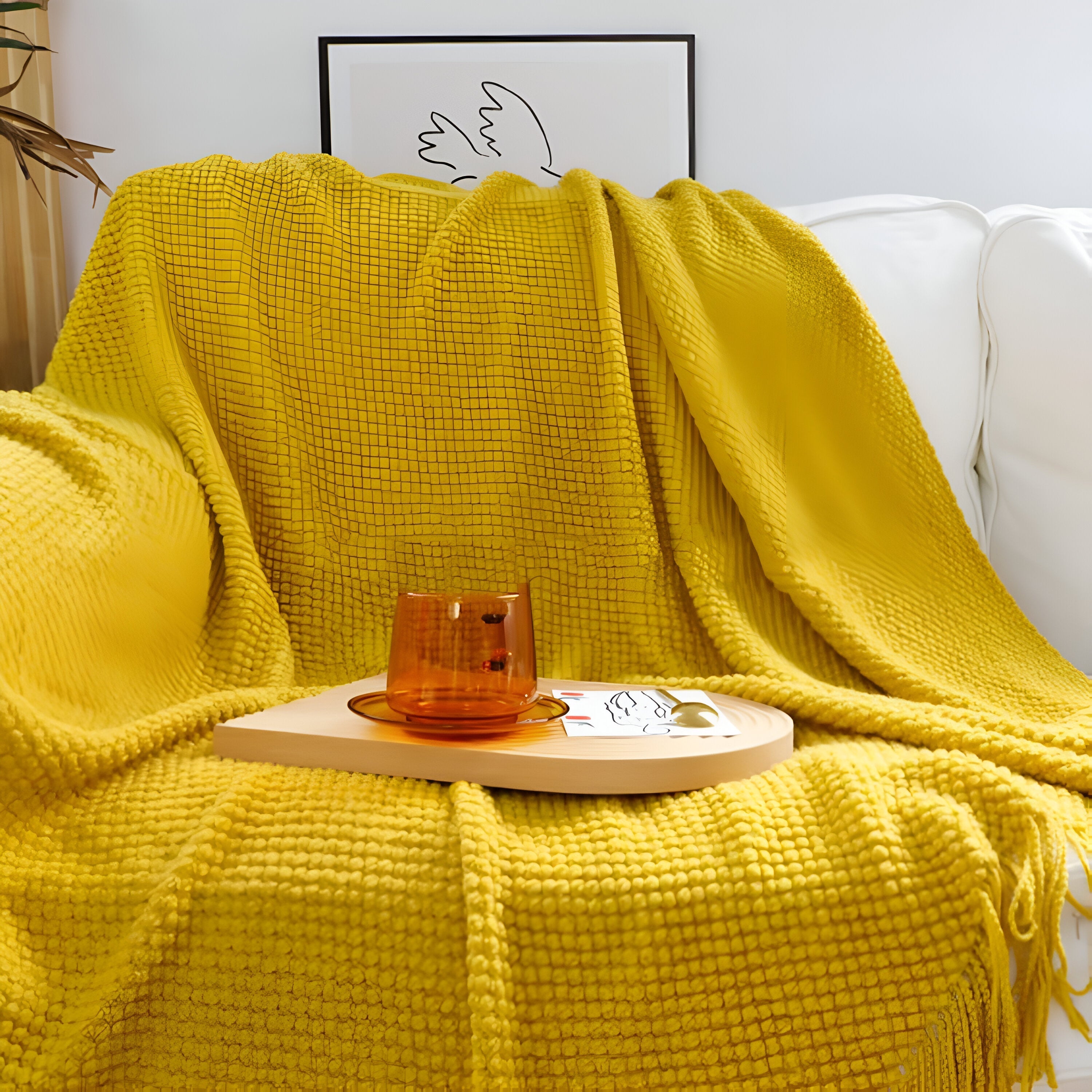 Yellow blanket draped over a white sofa with a small wooden table and candle on a light-colored wall background.
