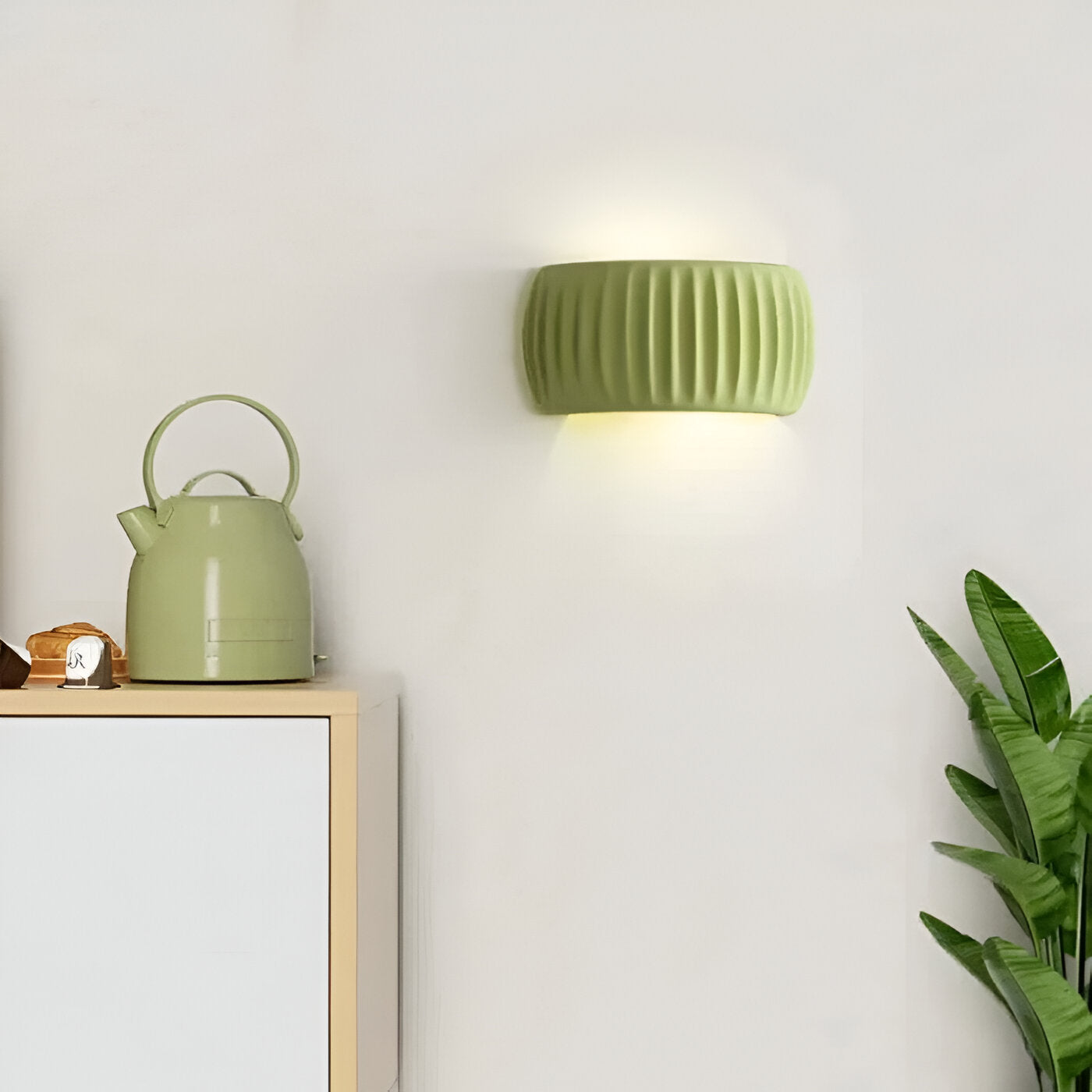 Green wall lamp and teapot on a wooden shelf against a white wall with a plant.
