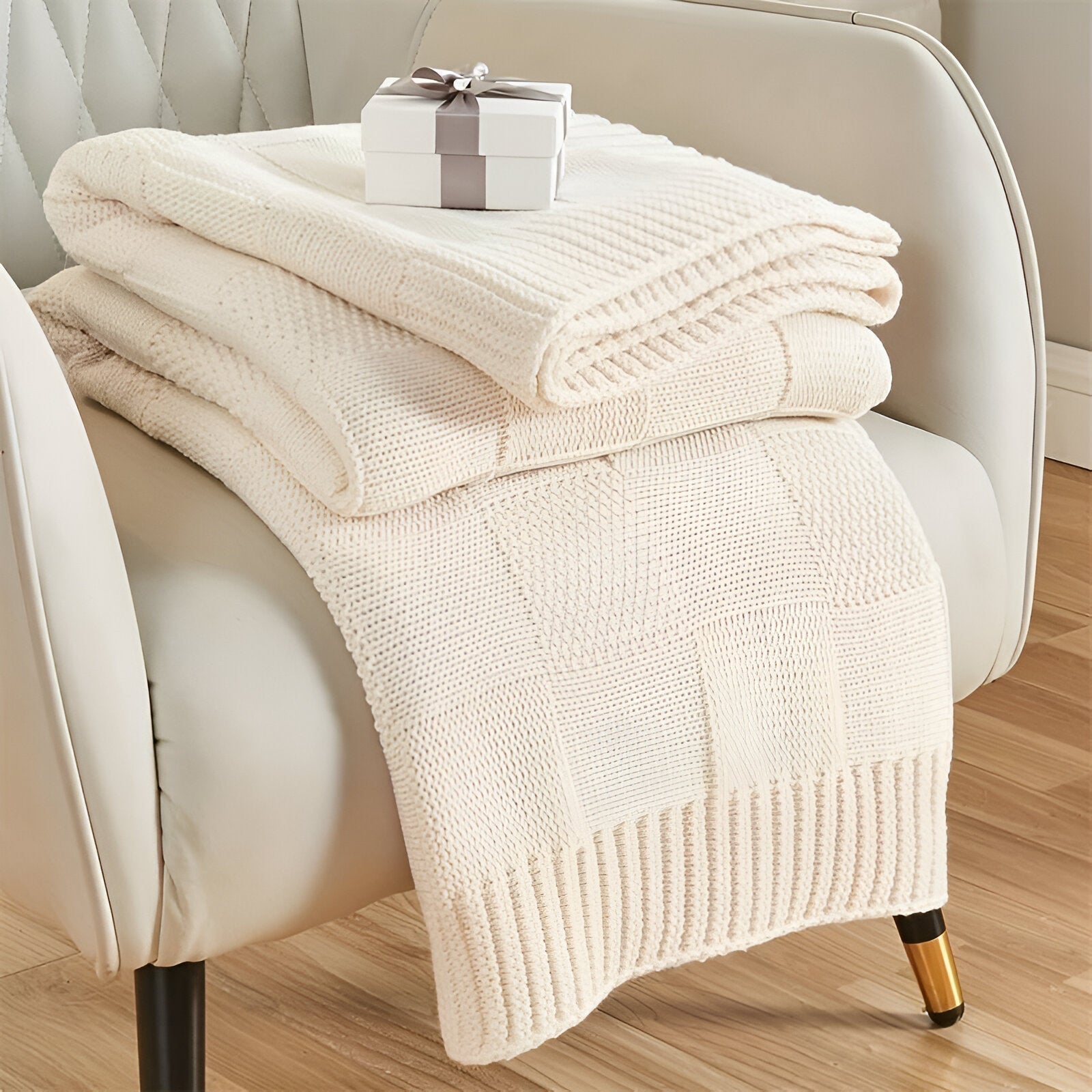 Folded beige blanket on a white armchair with a small gift box on top