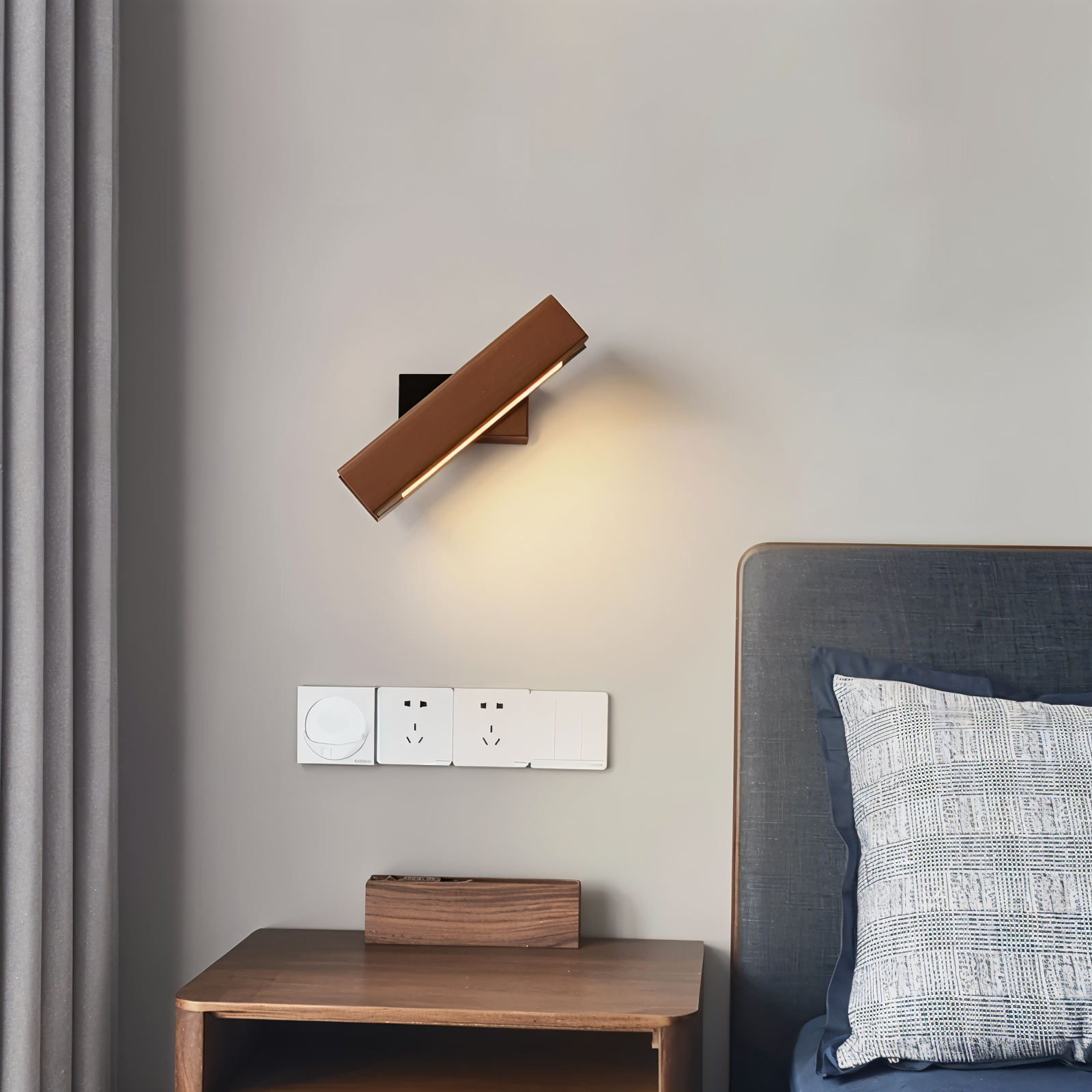 Modern wall lamp on a gray wall above a wooden nightstand with a bed and pillow in the background.