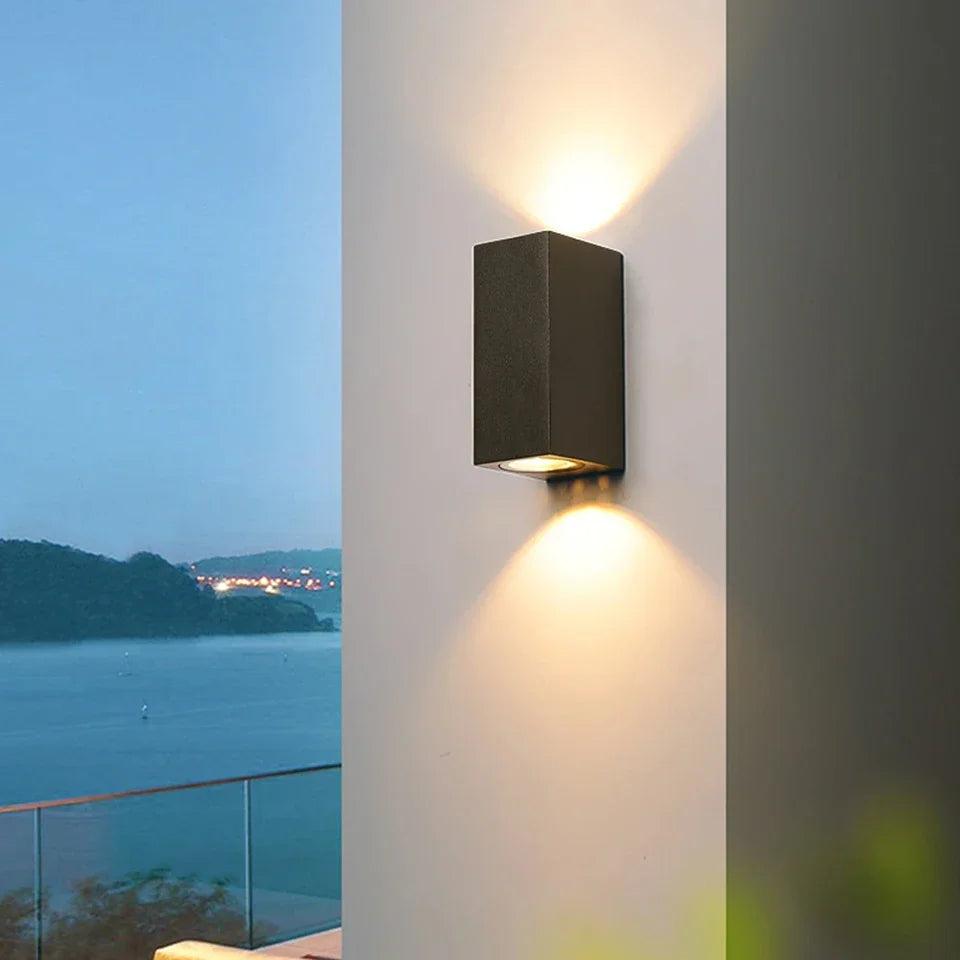 Wall-mounted outdoor light fixture on a building with a scenic view of water and lights in the distance.