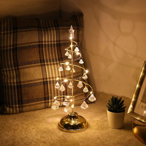Small decorative Christmas tree with lights on a table next to a plaid blanket and a plant.