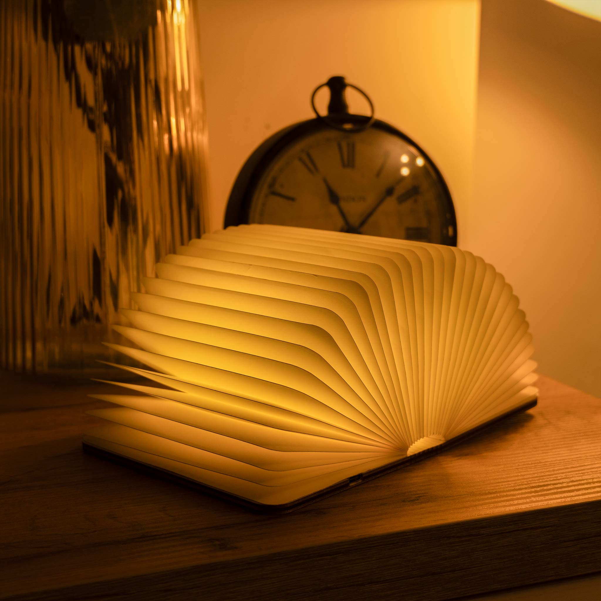 Book-shaped lamp emitting a warm glow on a wooden surface with a clock in the background.