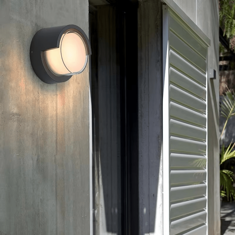 Round outdoor wall light fixture on a concrete wall with a corrugated metal shutter.