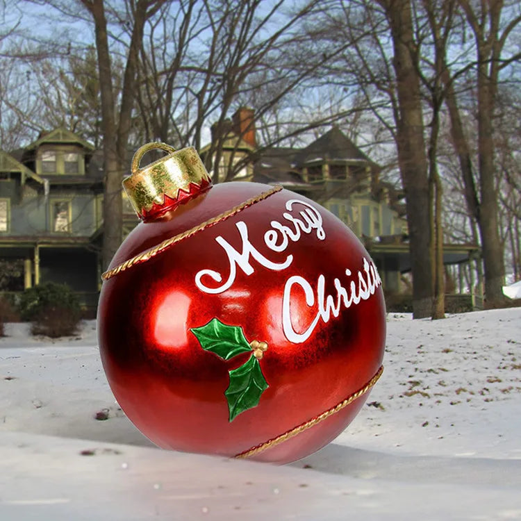 Red Christmas ornament with 'Merry Christmas' text and holly leaf in front of a snowy house.