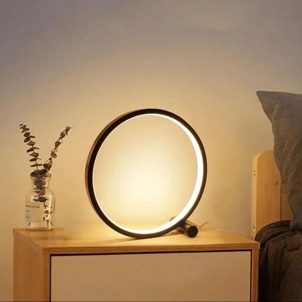 Round LED table lamp on a nightstand with a vase of eucalyptus and a bed in the background.