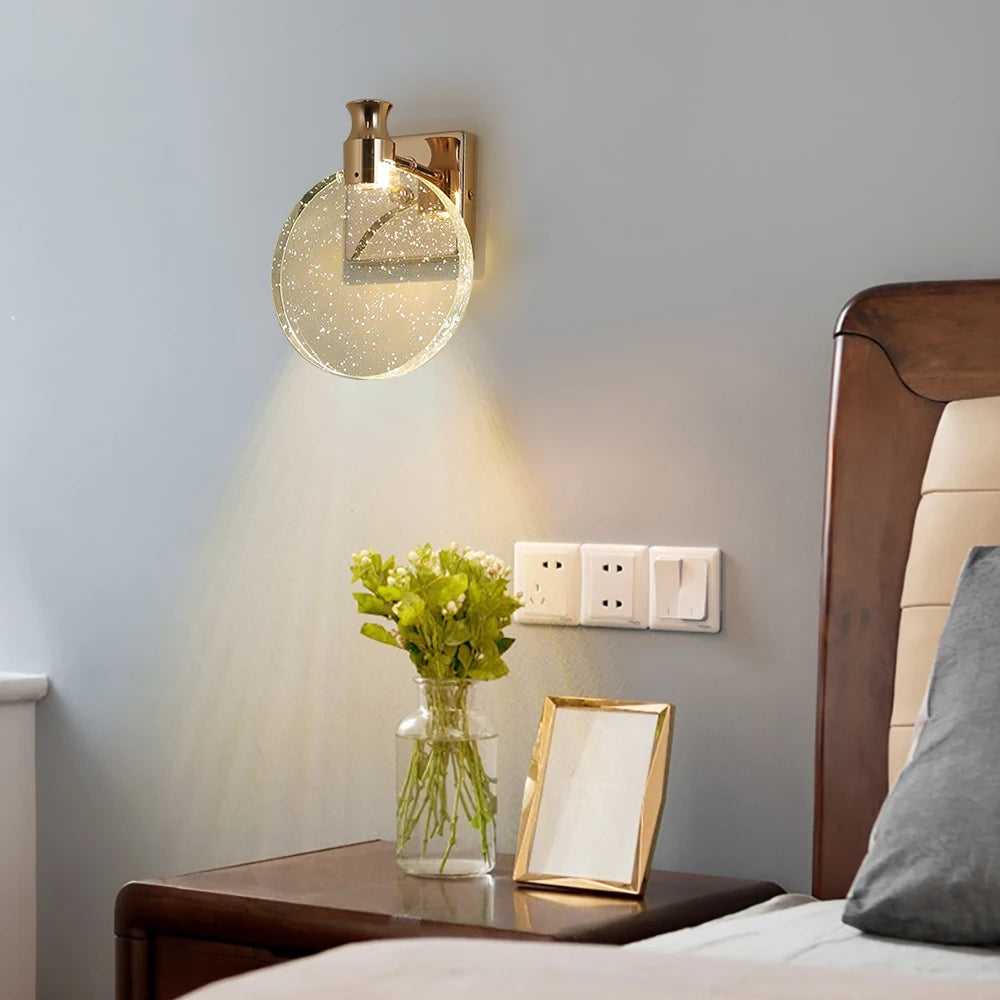 Gold wall sconce on a gray wall above a wooden nightstand with a vase of flowers and a mirror.