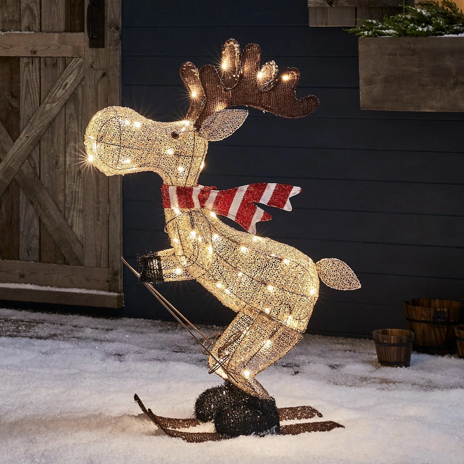 Decorative moose with lights and a red and white striped scarf in a snowy setting.